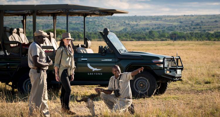 Safari guide points out wildlife signs to two guests beside a green open 4x4 in golden grasslands.