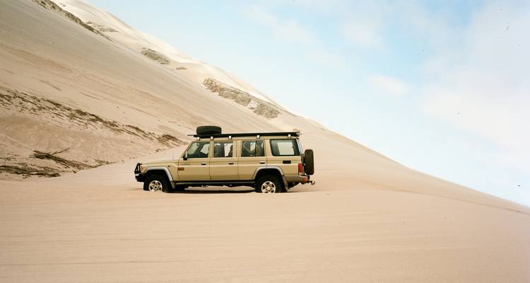 A 4x4 vehicle partially buried while driving across towering desert sand dunes under a blue sky.