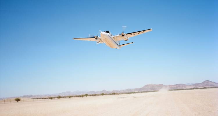 A small twin-engine aircraft takes off low over a dusty desert runway against a cloudless blue sky.