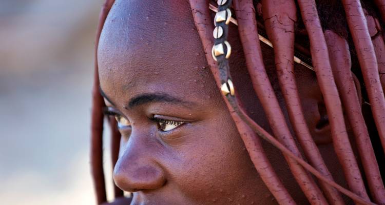 Close portrait of a Himba woman showing traditional ochre-covered braided hair decorated with shells.