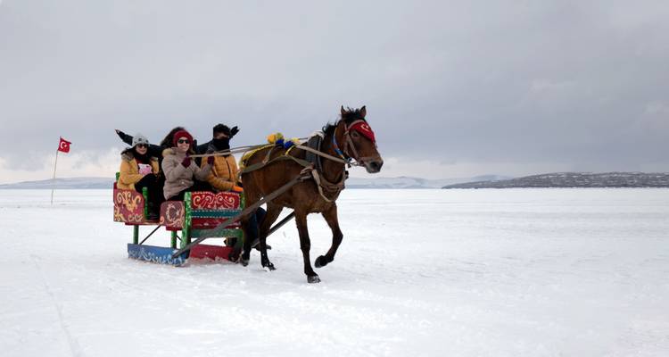 Traîneau tiré par des chevaux transportant des touristes emmitouflés à travers un vaste lac gelé avec des collines lointaines.