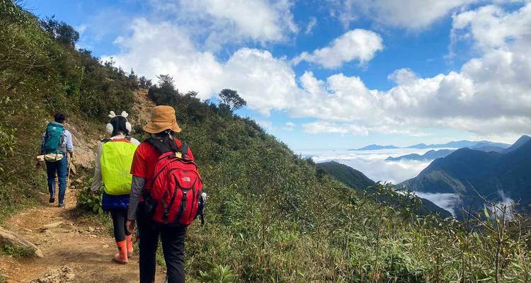 Un petit groupe de trekkers marche le long d'un sentier de montagne bordé de nuages sous un ciel bleu parsemé.