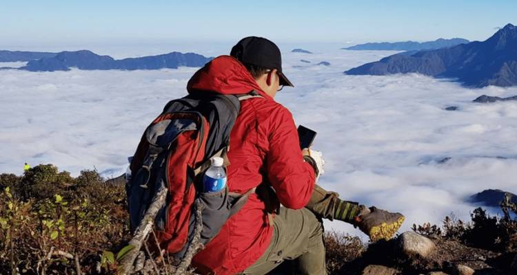 Un randonneur vêtu d'une veste rouge est assis sur un rebord rocheux au-dessus d'une couche de nuages blancs et consulte son téléphone.