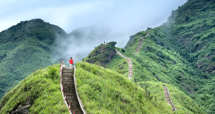 Un escalier abrupt serpente le long d'une crête d'un vert éclatant qui disparaît dans la brume, avec une silhouette solitaire vêtue de rouge qui gravit les marches.