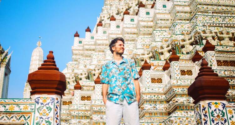Un jeune homme vêtu d'une chemise à motifs floraux pose devant les tours richement décorées d'un temple thaïlandais au bord d'une rivière sous un ciel bleu dégagé.