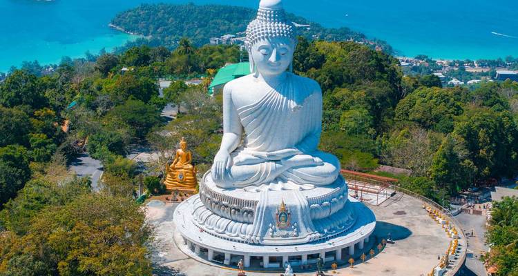 Une grande statue de Bouddha blanche est perchée au sommet d'une colline verte dominant une mer turquoise et des îles.