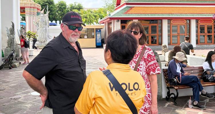 Un guide touristique discute avec des visiteurs à l'extérieur d'un complexe de temples à Bangkok par une journée ensoleillée.