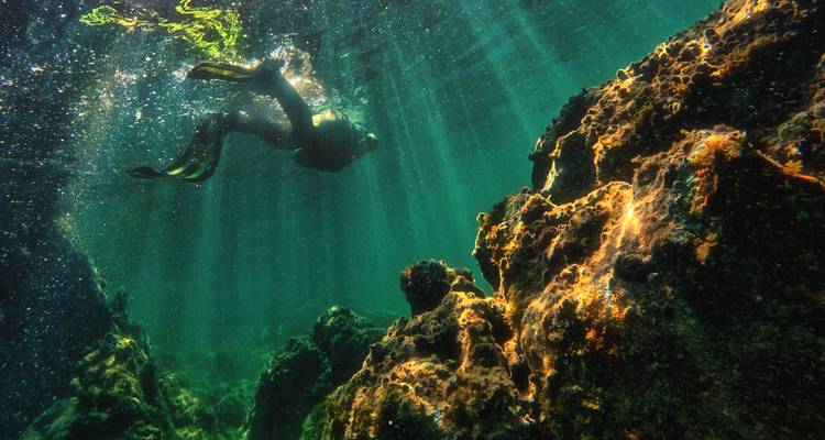 Un plongeur en apnée glisse entre des rochers lumineux dans une eau turquoise cristalline avec des rayons de soleil qui filtrent d'en haut.
