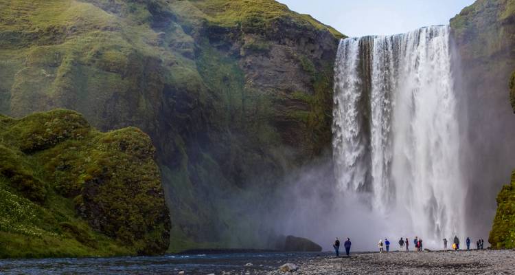 Le puissant rideau de Skogafoss gronde en se transformant en brume tandis que les visiteurs se tiennent rétrécis à sa base.