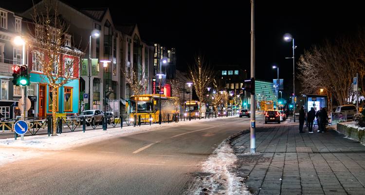 Rue de Reykjavik saupoudrée de neige la nuit, illuminée par des devantures de magasins colorées, des autobus et des lampadaires.