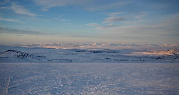 Une vaste étendue sauvage couverte de neige s'étend jusqu'aux montagnes aux teintes roses lointaines sous un ciel pastel.