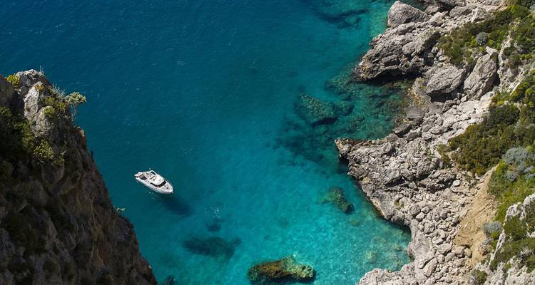 Vue de dessus d'un petit bateau blanc flottant dans une crique turquoise aux eaux cristallines, entourée de falaises rocheuses.