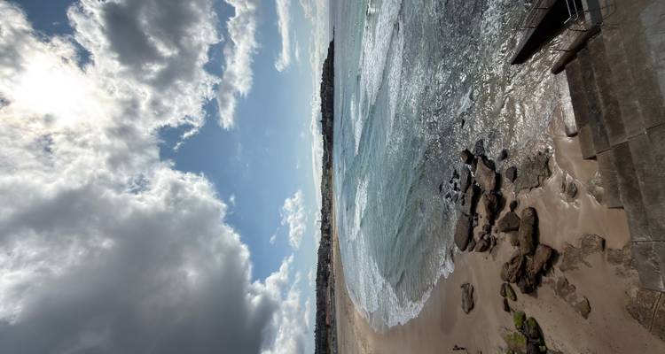 Una bahía arenosa y extensa con olas suaves bajo un mosaico de sol y nubes.
