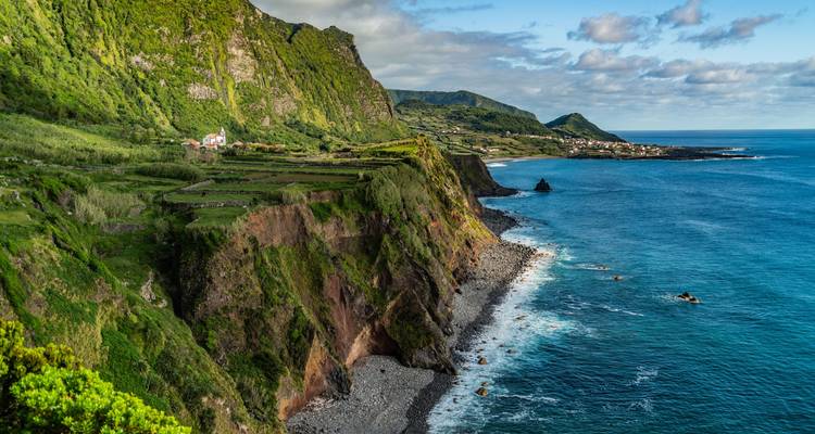 Des falaises vertes abruptes descendent vers un rivage rocheux avec un village lointain et une mer bleue.