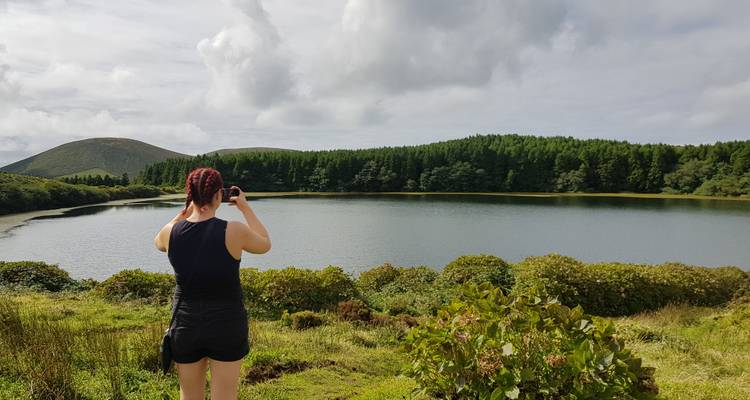Un voyageur photographie un lac serein entouré de forêt et d'arbustes sous un ciel nuageux.