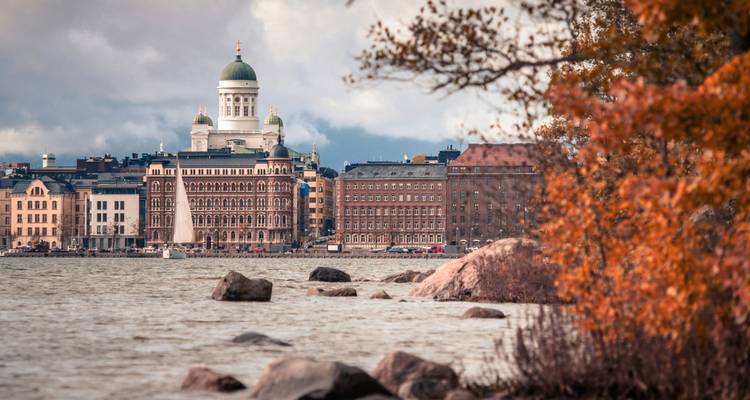 La Catedral de Helsinki con su cúpula blanca se alza sobre los edificios históricos del paseo marítimo, enmarcada por hojas de otoño anaranjadas y un cielo nublado.