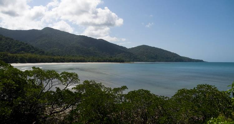 Plage courbe immaculée bordée par la forêt tropicale avec des montagnes s'élevant derrière sous un ciel lumineux.