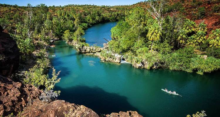 Uitzicht van bovenaf op een slingerende turquoise rivier omgeven door rode rotsachtige kliffen en dicht oerwoud, met kajakers die eronder peddelen.