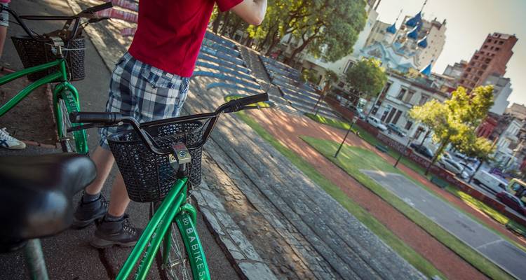 Un cycliste en pause avec un vélo de location vert surplombant un parc urbain ensoleillé et l'architecture de la ville