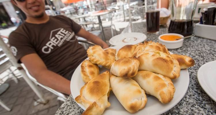 Un plato lleno de empanadas recién horneadas y doradas en una mesa de café al aire libre con vasos de cerveza.