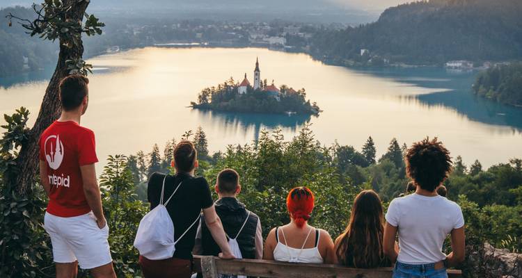 Travellers sit on a bench overlooking serene Lake Bled with its small island church in the golden evening light.