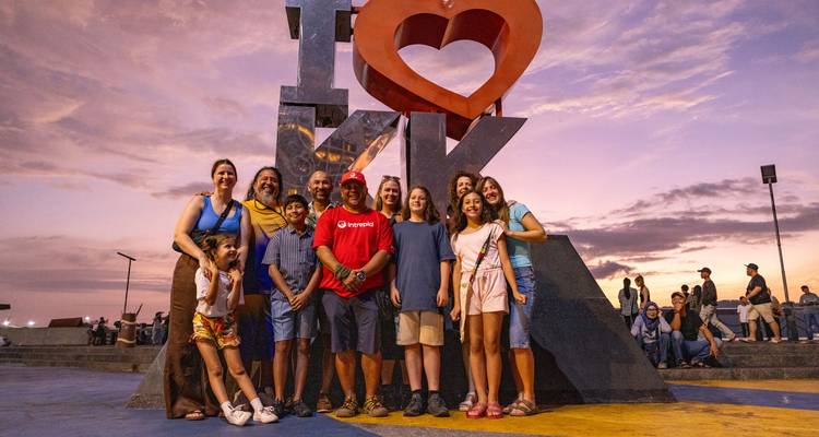 Un gran grupo de viajeros sonrientes posan frente a una escultura de "I ❤ KK" con una puesta de sol colorida de fondo.