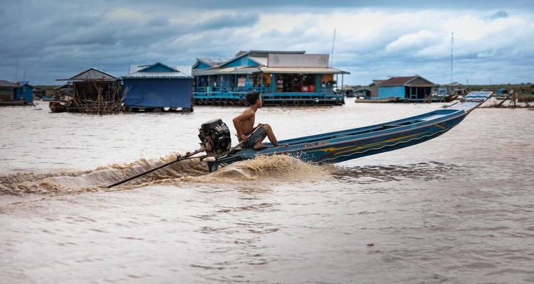 Un hombre en un barco de cola larga en un lago, con casas flotantes en el fondo.