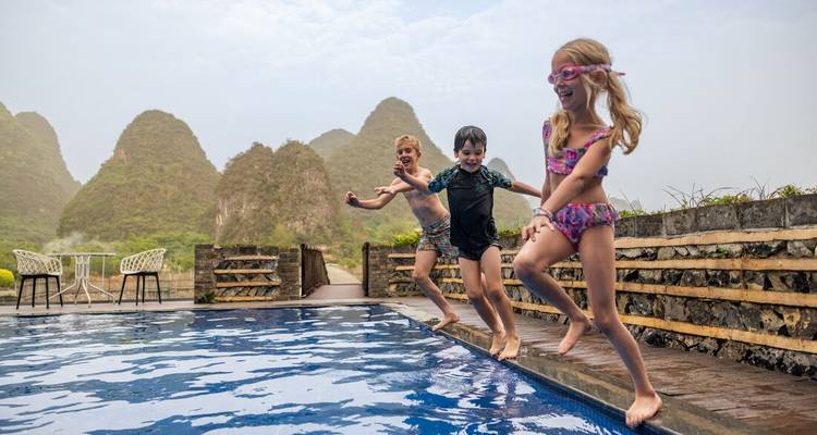 Three children jumping into a pool with karst hills in the background.