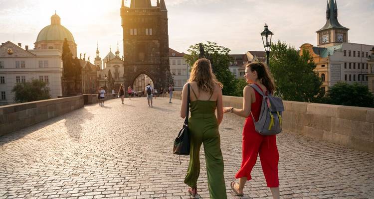 Twee vrouwen wandelen over de Karelsbrug in Praag, gebaad in gouden avondlicht, richting de gotische toren.