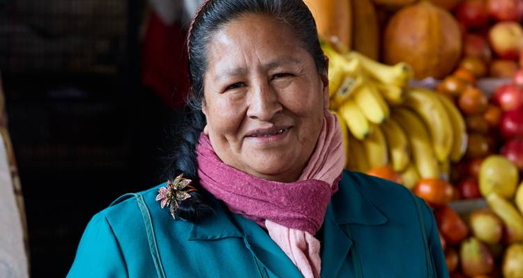 A person smiling in a market setting with fruits in the background.