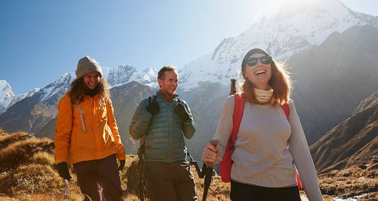 Drie glimlachende wandelaars lopen door een zonnige alpine weide met dramatische sneeuwtoppen achter hen.