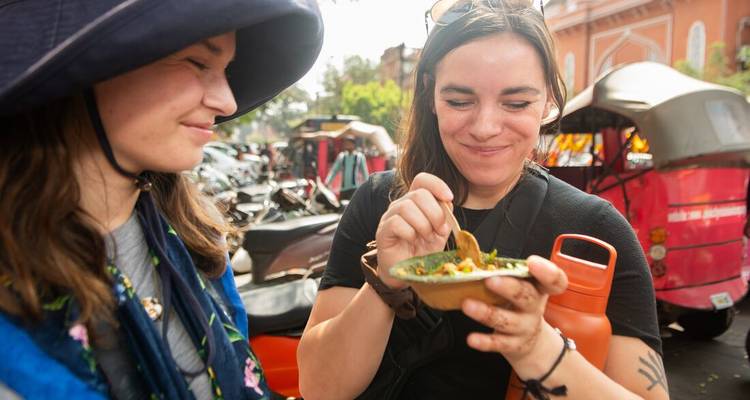 Two travelers sample flavorful street food from a small bowl while exploring a bustling Indian market.