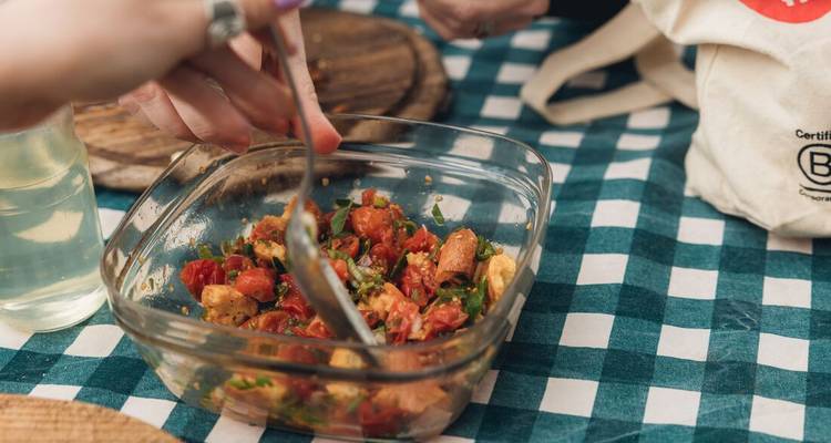 Des mains mélangent une salade fraîche de tomates, basilic et pain dans un plat en verre sur une nappe à carreaux.