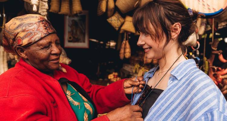 Local vendor fastens a beaded necklace on a smiling traveller at a bustling market stall.
