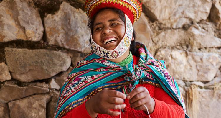 Retrato en primer plano de una mujer andina sonriente que lleva un sombrero bordado colorido y un chal contra una pared de piedra.