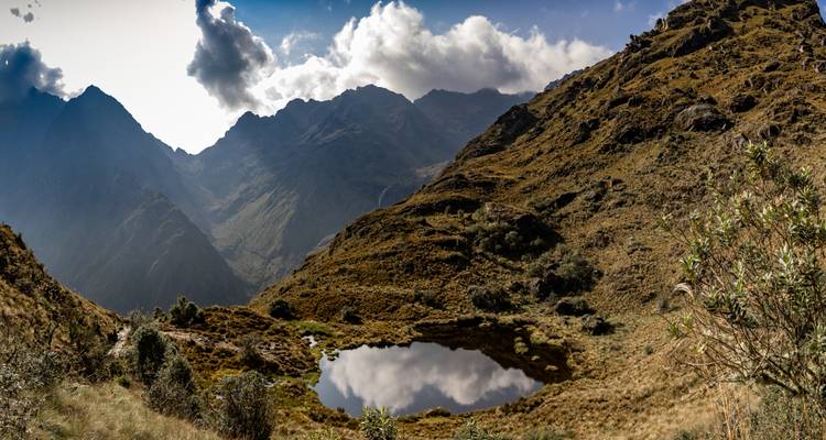 Amplio paisaje alpino con cimas dentadas, un estanque reflectante y praderas onduladas marrones bajo un cielo brillante.