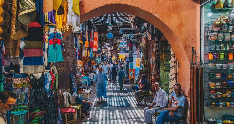 Ruelle animée du souk de Marrakech bordée de textiles colorés, de poteries et de habitants locaux sous un toit en treillis.