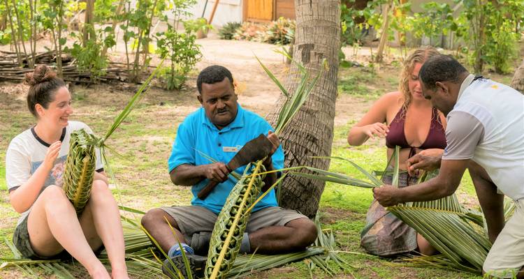 Kleine Gruppe sitzt im Gras und flicht Palmwedel-Handwerk in einem fidschianischen Dorfumfeld