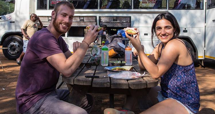 Deux personnes dégustant un repas à l'extérieur, près d'un véhicule de safari.