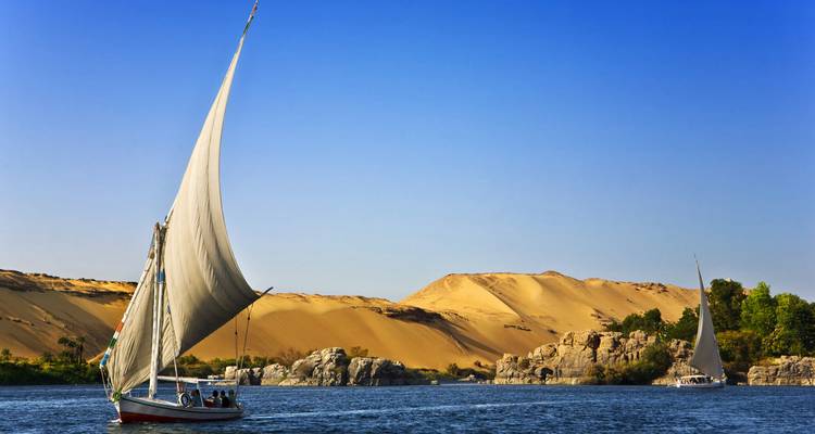 Traditionele felucca met hoog zeil glijdt over de blauwe Nijl tegen de achtergrond van gouden woestijnduinen.