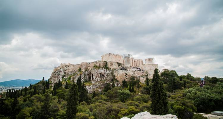 Vista lejana de la Acrópolis encaramada en lo alto de una colina rocosa rodeada de árboles verdes bajo nubes amenazantes.