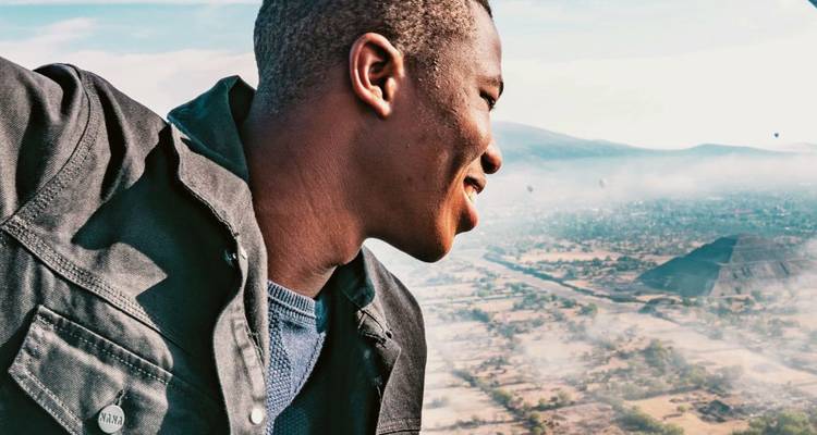 Smiling man leaning from a hot-air balloon basket above misty plains and Teotihuacán pyramids.