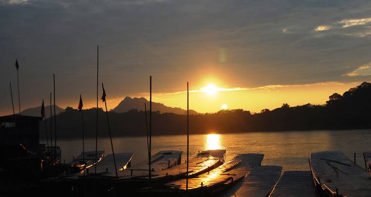 Amanecer dorado sobre un río tranquilo con siluetas de barcas de cola larga amarradas y montañas lejanas