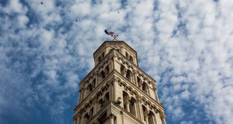 Vista hacia arriba de la torre campanario de la catedral de Split bajo un cielo salpicado de nubes con patrones y pájaros.