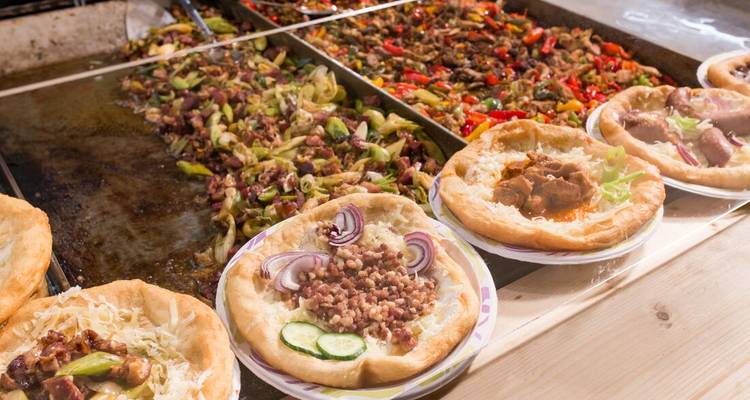 Hungarian street food langos topped with meat, onions and cheese sizzling beside colourful peppers.