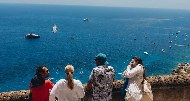 Small group of travellers admiring a panoramic blue-sea vista dotted with yachts from a cliffside viewpoint