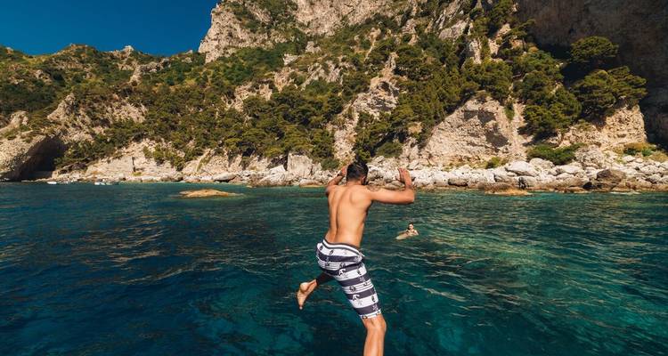 Man mid-air jumping off a boat into clear turquoise water near rugged cliffs