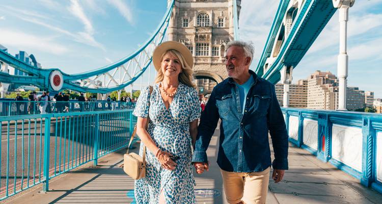 An older couple holding hands walks across the vibrant blue deck of Tower Bridge in London.