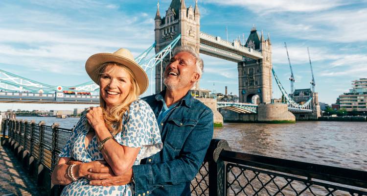 Laughing couple embraces on a riverside promenade with Tower Bridge arching behind them.