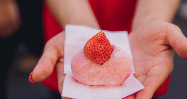 Une main tenant un dessert de mochi rose surmonté d'une fraise fraîche sur un emballage en papier.